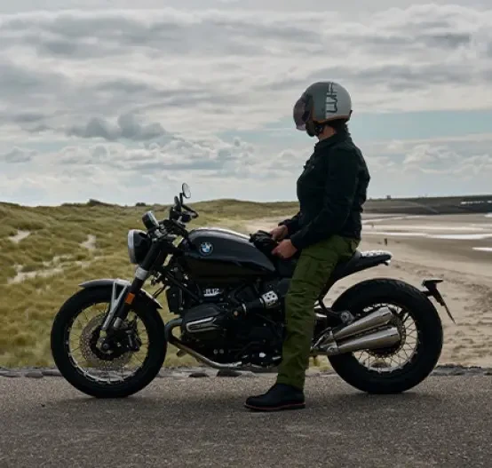 Static left side image of BMW Motorrad R 12 nineT Option 719 in Black Storm Metallic colourway, with female rider sitting on the motorcycle, beach background